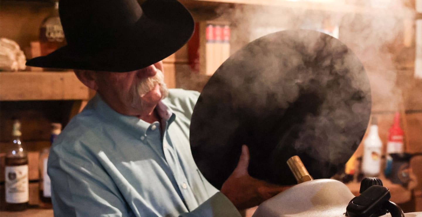 A cowboy wearing a black hat, holding a black hat at a steam machine, heating the hat with steam.